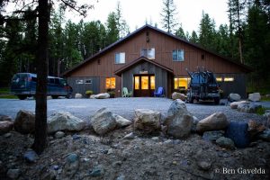 Brown lodge at a Glacier National Park campground with a gravel lot, parked vans, bike rack, and pine forest backdrop.