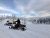 Snowmobiles sit on a snow-covered ridge at Whitefish Bike Retreat in Glacier National Park, with frosted evergreen trees and distant snowbound peaks.