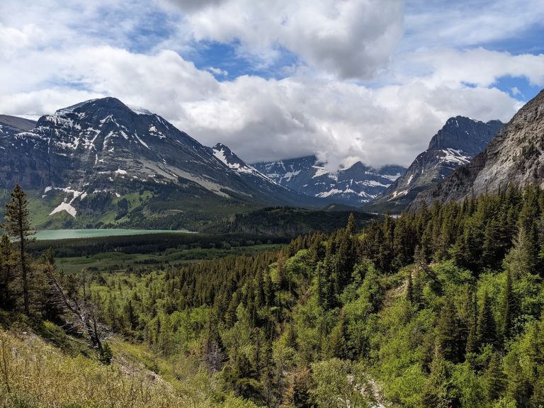 Apikuni Falls overlook at Glacier National Park, with snow-dusted peaks, lush forest, and a turquoise lake in a dramatic valley.