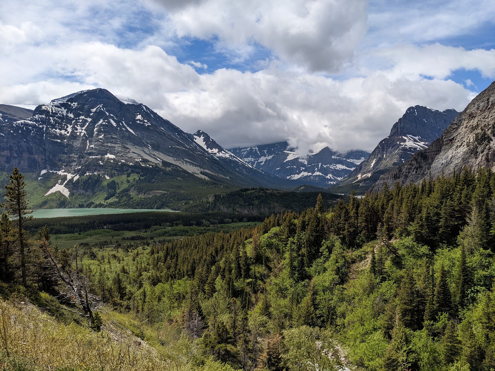 Apikuni Falls overlook at Glacier National Park, with snow-dusted peaks, lush forest, and a turquoise lake in a dramatic valley.