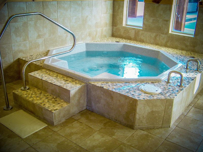 Indoors, a hexagonal hot tub with a mosaic tiled edge, metal handrails, beige walls, and two windows in Yellowstone National Park.