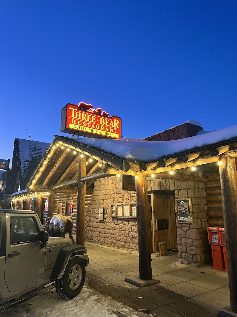 Snowy log exterior with warm string lights, a Jeep parked outside, and a rustic restaurant entrance in Yellowstone National Park.