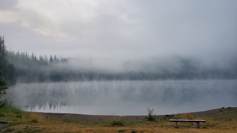 Mist over a quiet lake at dawn in Glacier National Park, pine shoreline, calm water, and a bench.