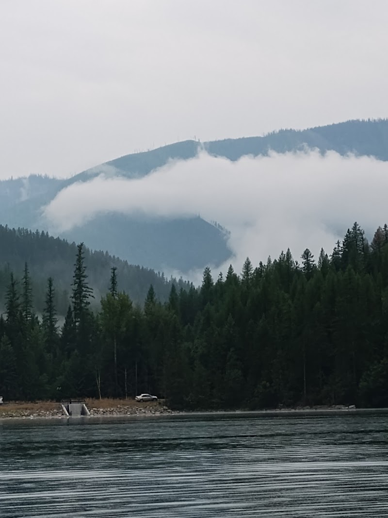 Misty pine-covered mountains loom over a quiet lake in Glacier National Park, with a gravel shore and a white car nearby.