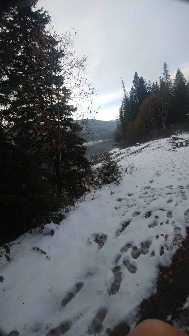 Snowy riverside trail in Glacier National Park with footprints, evergreen trees, and a distant lake along a forested shoreline.