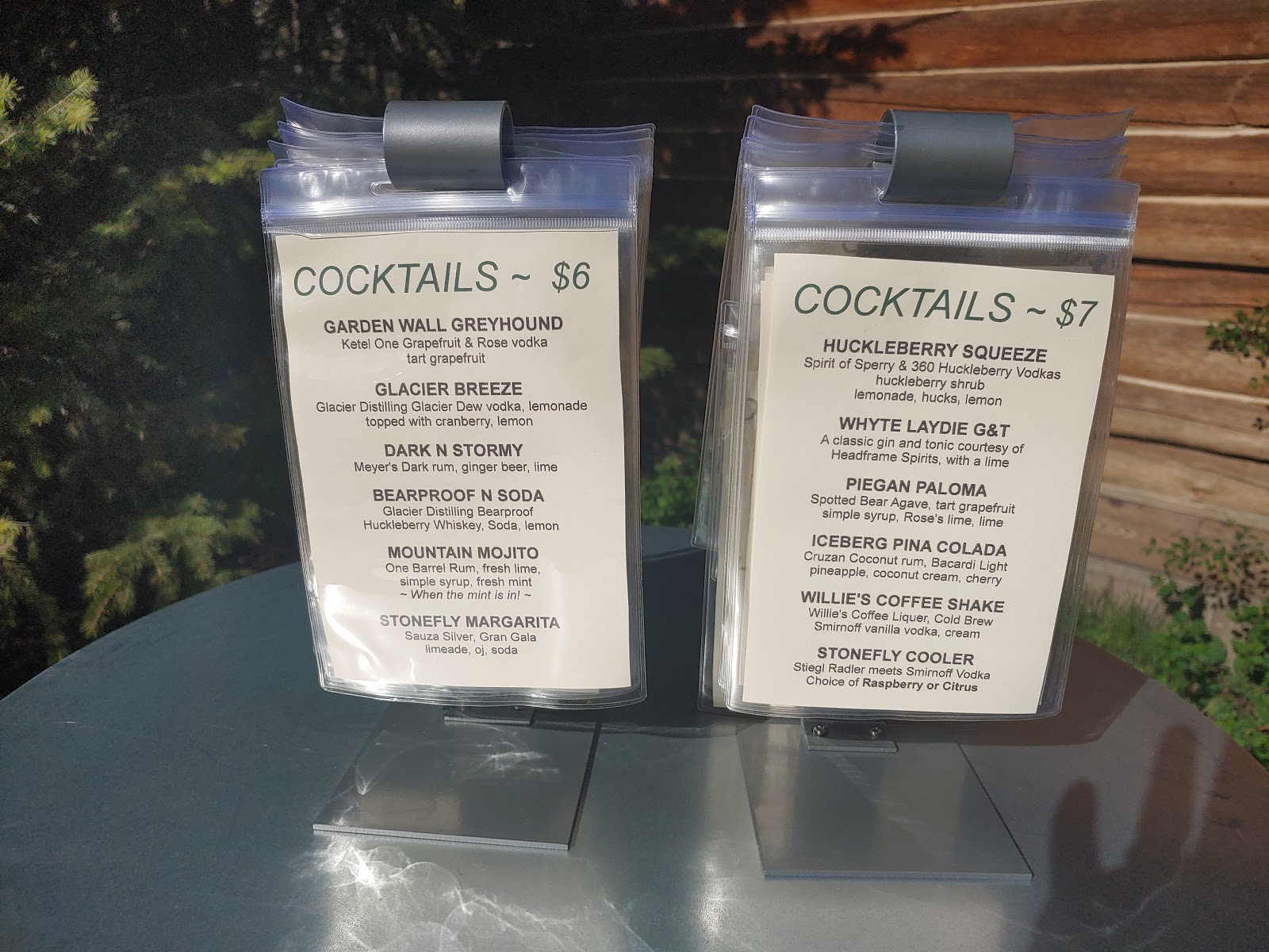 Two clear plastic menu holders display cocktails on a round metal table, with a wooden wall backdrop in Glacier National Park.