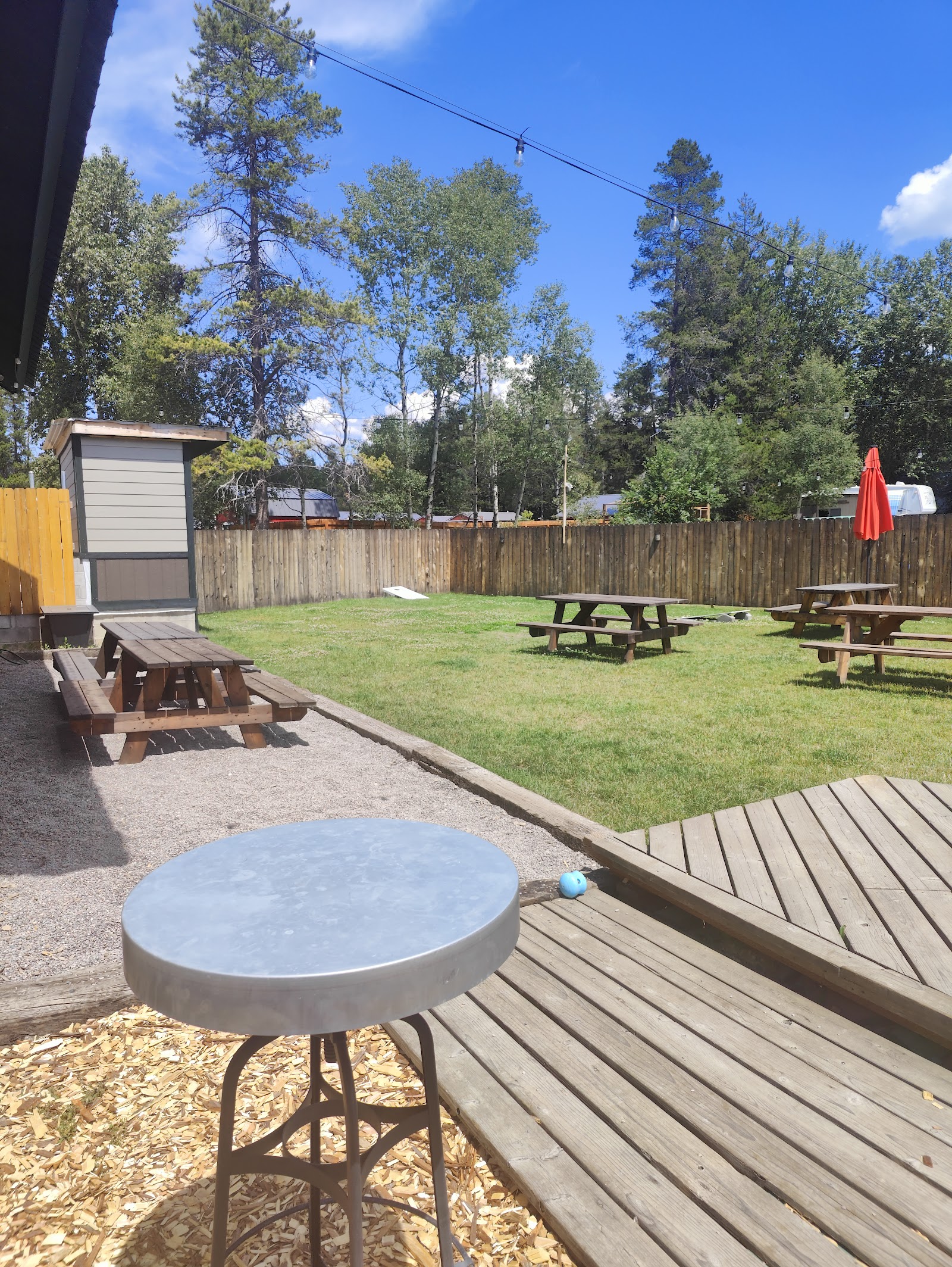 Sunny outdoor patio with wooden picnic tables, a red umbrella, string lights, and tall trees in Glacier National Park.