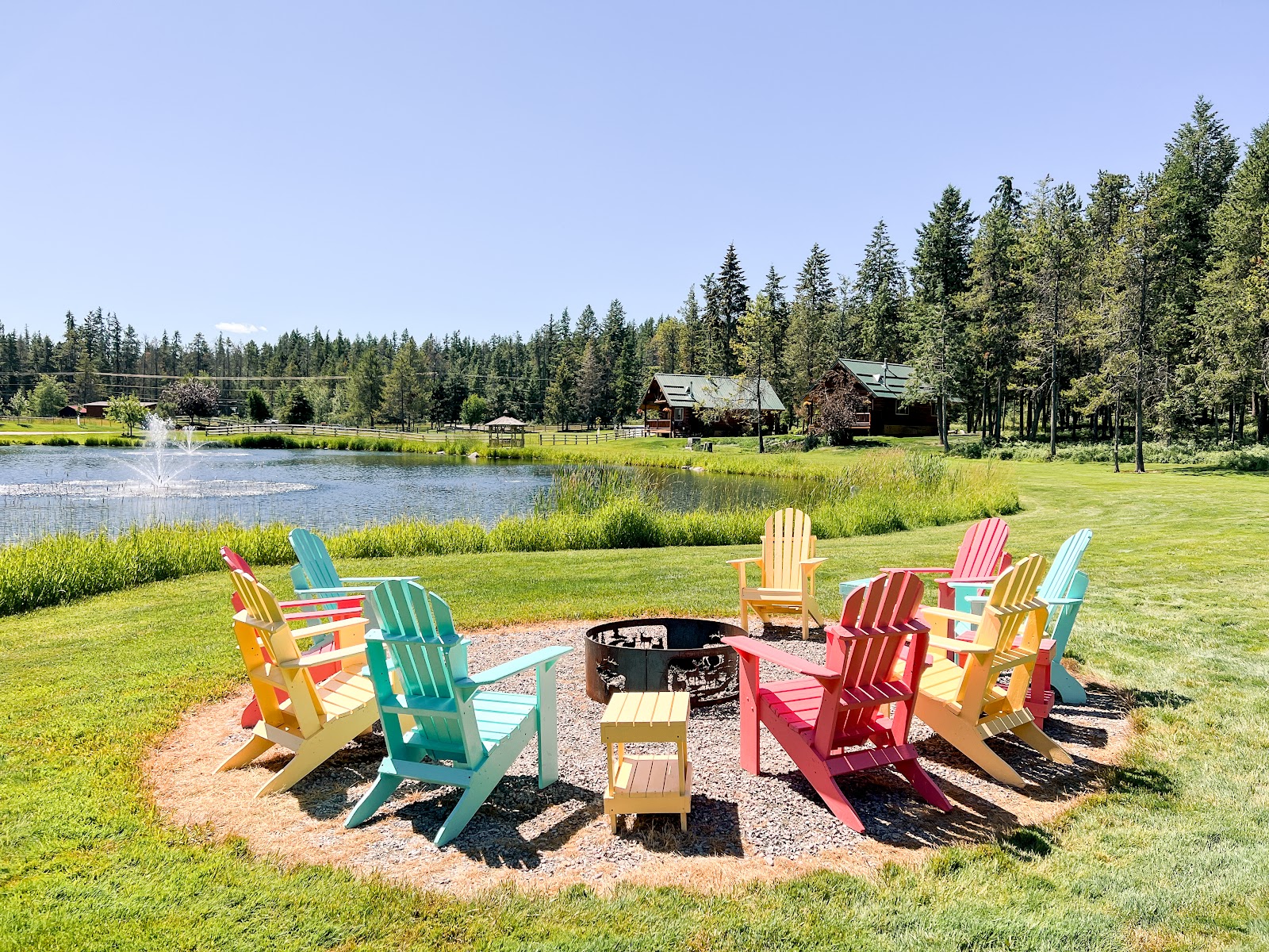 Colorful Adirondack chairs surround a fire pit beside a pond at a Glacier National Park lodging area.