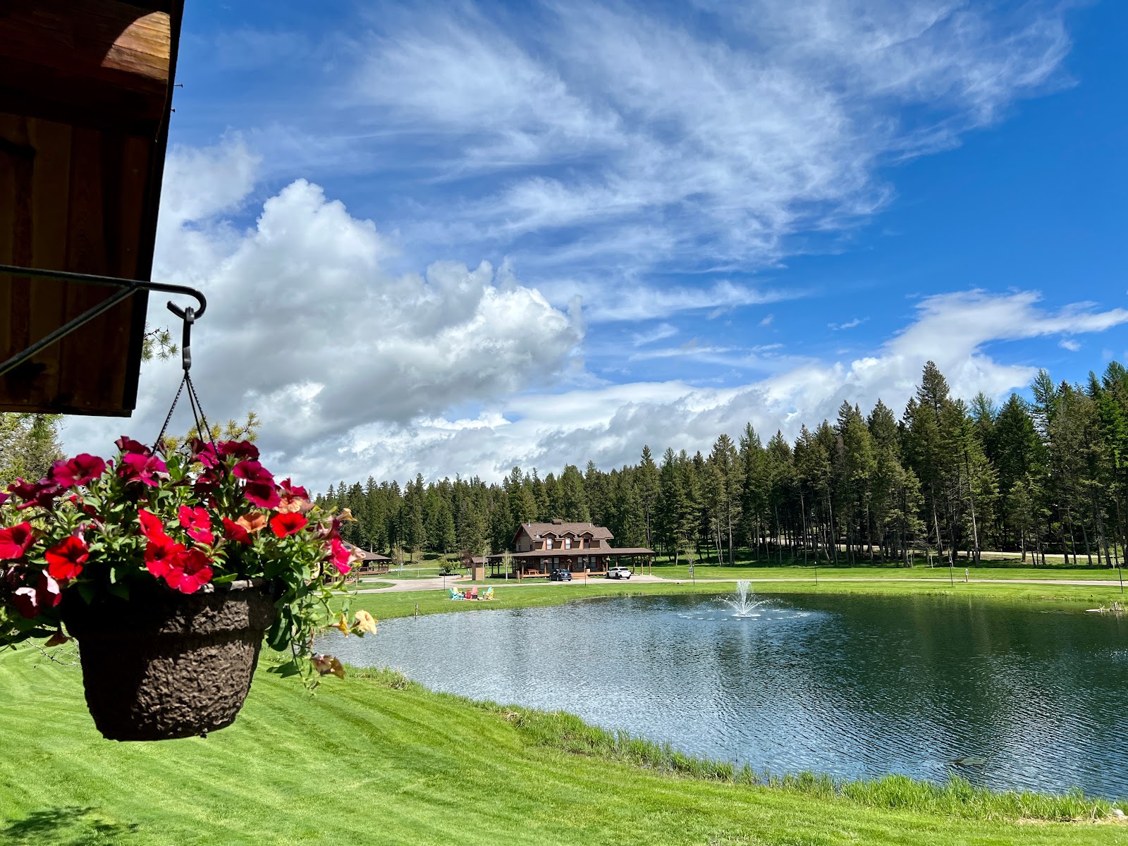 Glacier National Park pond with a fountain, a wooden lodge on the shore, and pine trees in the background under a bright blue sky.