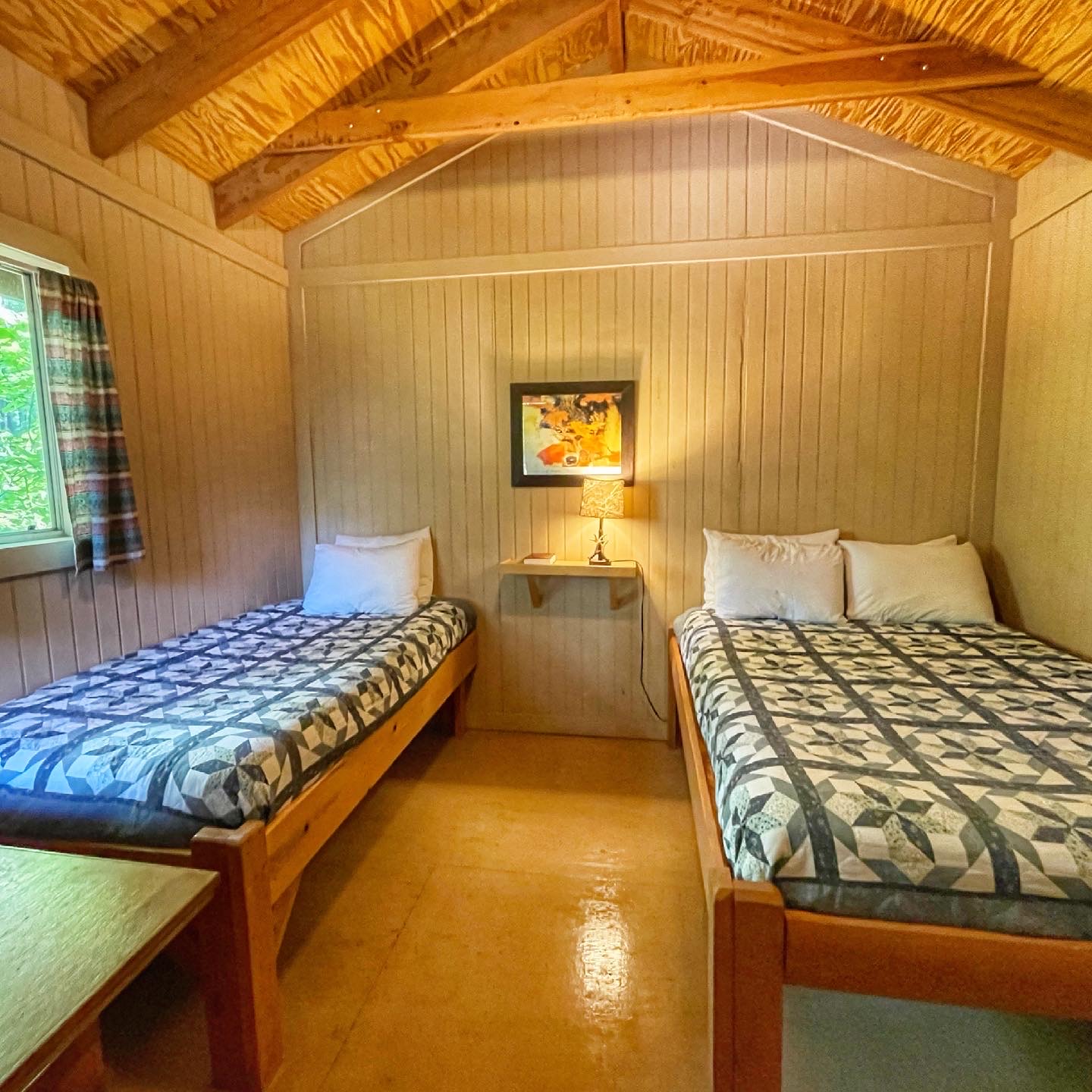 Timber-paneled cabin with two twin beds in patchwork quilts, a small nightstand lamp, and a window with plaid curtains in Glacier National Park.