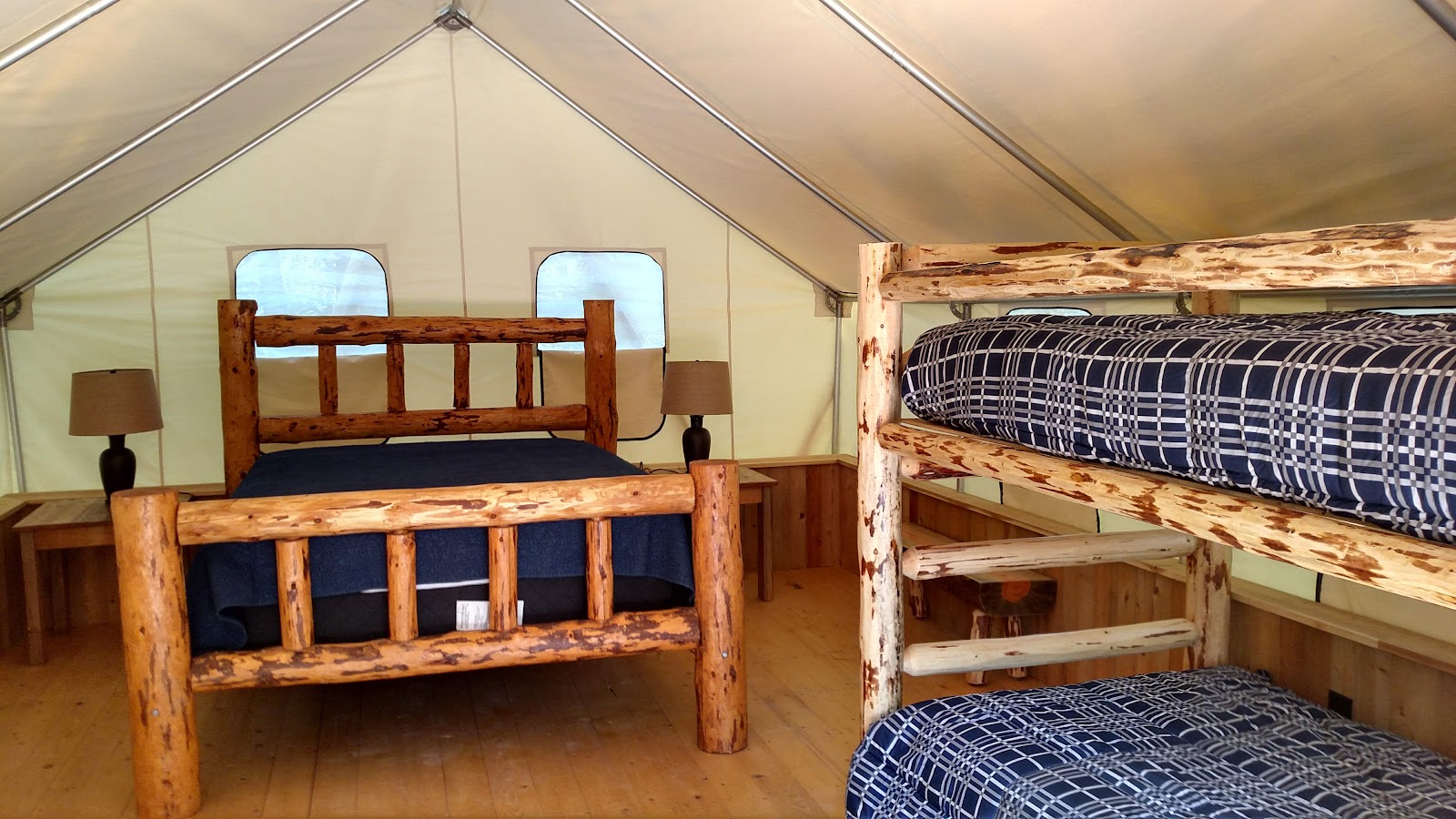 Rustic tent cabin in Glacier National Park showing a large log bed, a bunk bed, plaid navy bedding, and side lamps.