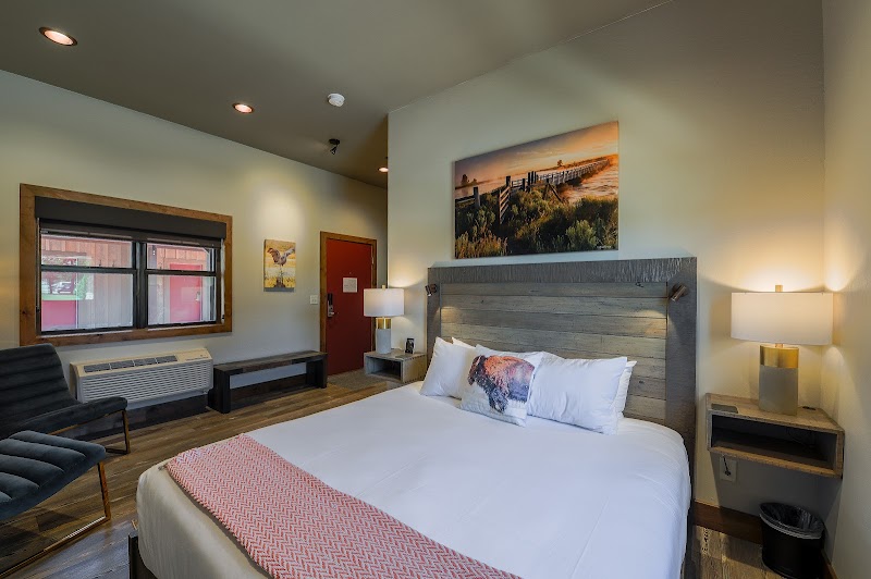 Cozy room with a large bed, grey wooden headboard, two nightstands, lamps, a window bench, and landscape art—Yellowstone National Park.