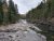 Slate-gray rock ledges and a shallow, winding river run through a rocky shore flanked by evergreen trees in Glacier National Park.