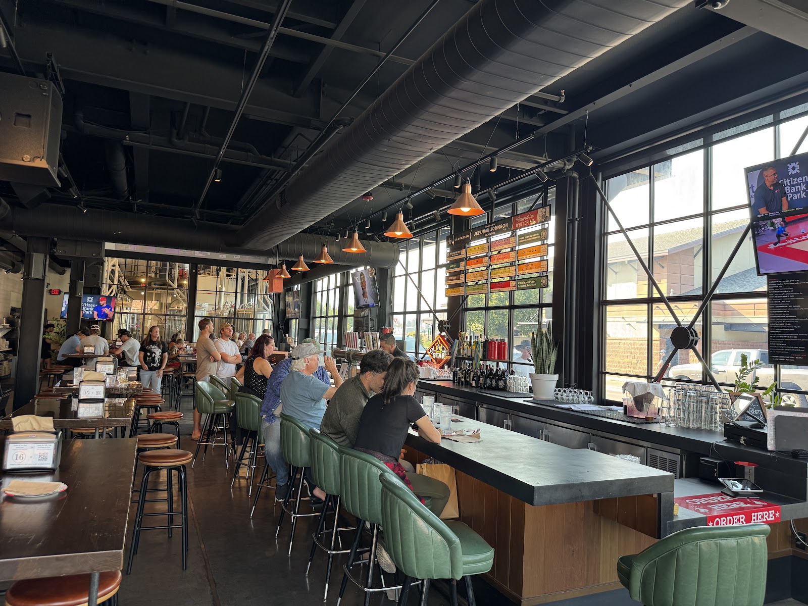 Industrial brewpub interior in Glacier National Park with a long bar, green stools, orange pendant lights, and large windows.