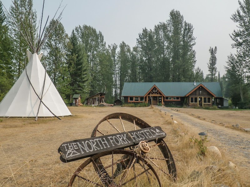 Rustic lodge and teepee at Glacier National Park along a dirt road near pine trees and a log pavilion.