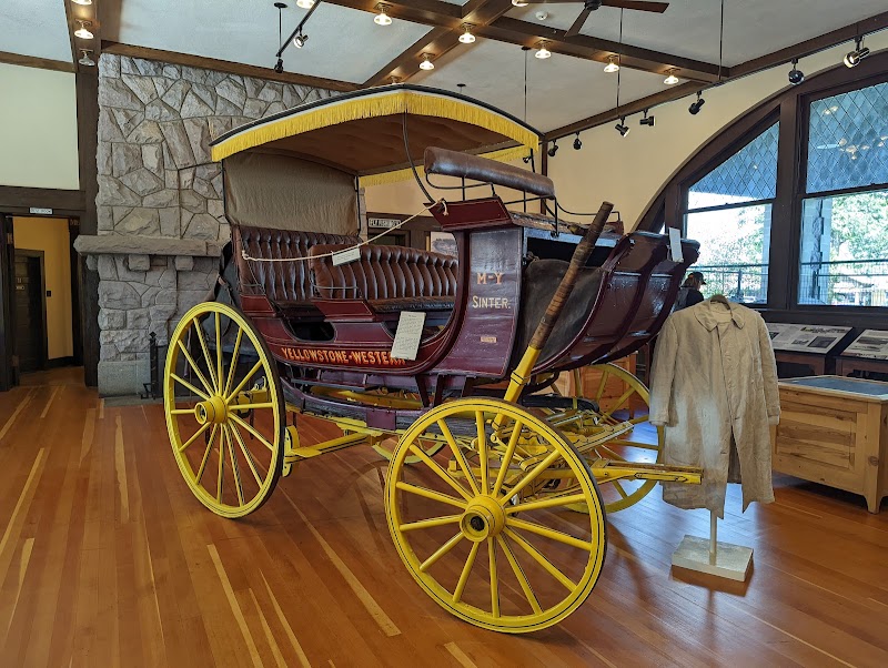 Historic stagecoach with yellow wheels and a canopy sits in a Yellowstone National Park museum, showcasing leather seats and a coat on a stand.