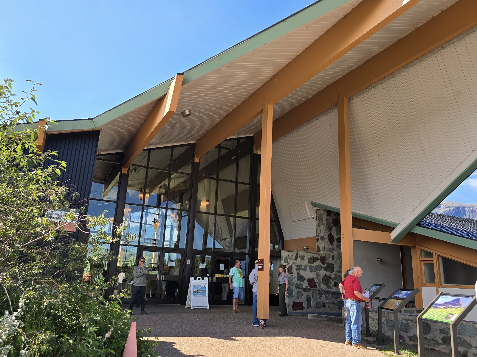 Glacier National Park visitor center with angular roof, orange posts, glass façade, and visitors viewing displays outside.