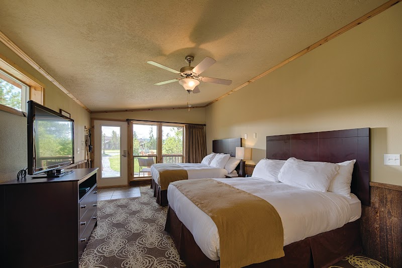 Two queen beds with white linens and tan throws, a ceiling fan, TV on a dark dresser, and a sliding door to a balcony in Yellowstone National Park.