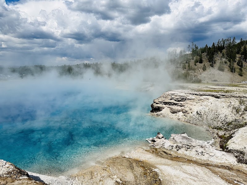 Grand Prismatic Spring glows blue in Yellowstone as steam drifts over mineral-rich edges and a forested bluff.