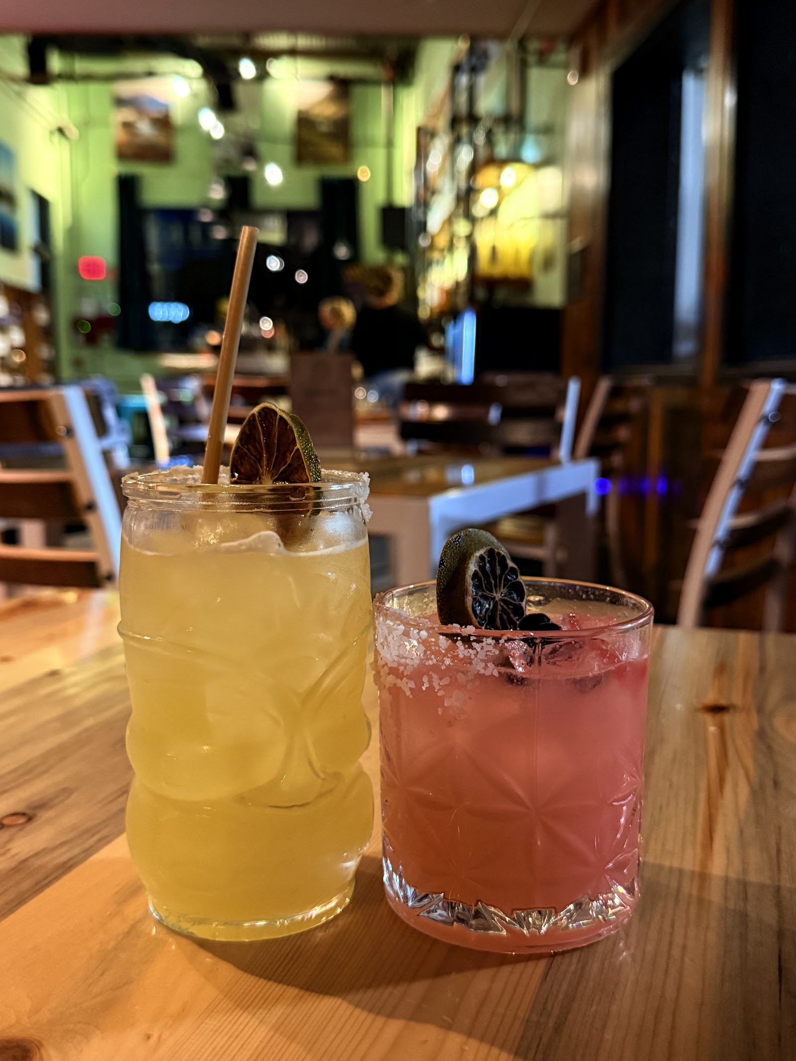 Inside a Glacier National Park bar, two colorful cocktails sit on a wooden table with ice and garnishes.