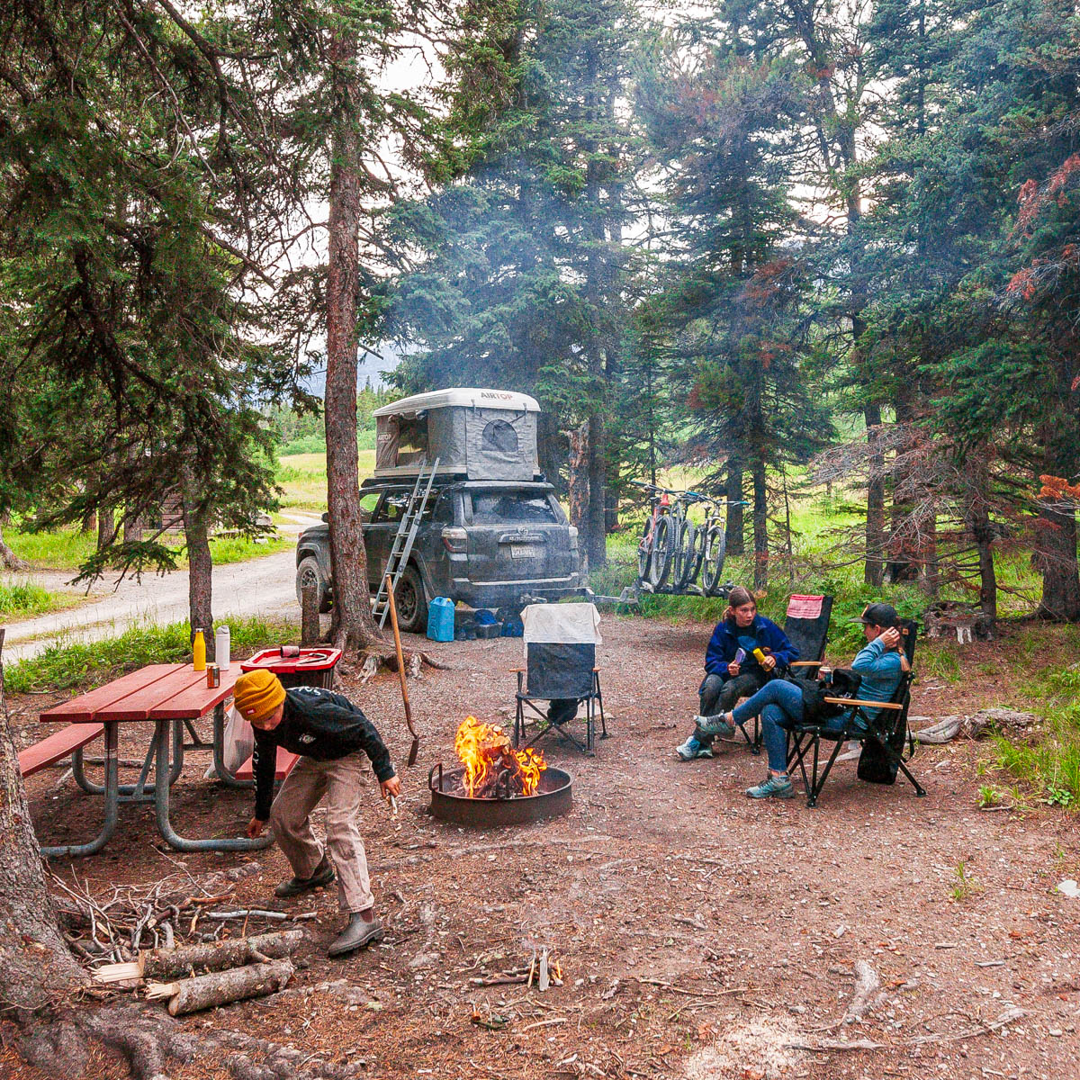 Glacier National Park campground with a car and rooftop tent, ladder, bikes, and people gathered around a campfire.