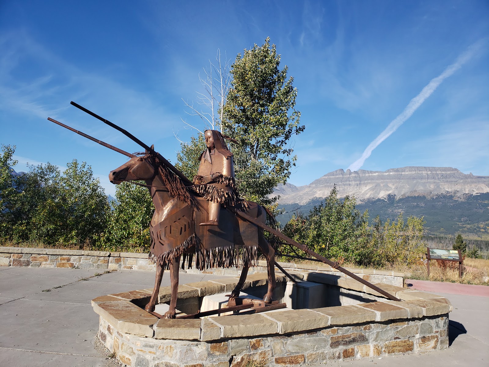 Rusty metal sculpture of a rider on a horse with spears, set in a stone platform, with Glacier National Park mountains.