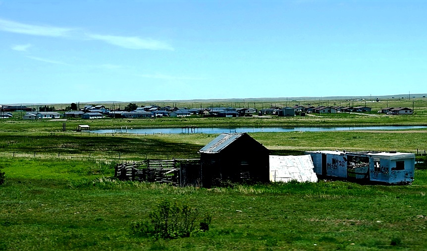 Rural scene in Glacier National Park: weathered shed and trailer beside a grassy field with a pond and distant houses.