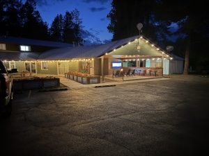 Cozy lodge with string lights, open-air bar and seating, flower planters, in Glacier National Park at night.
