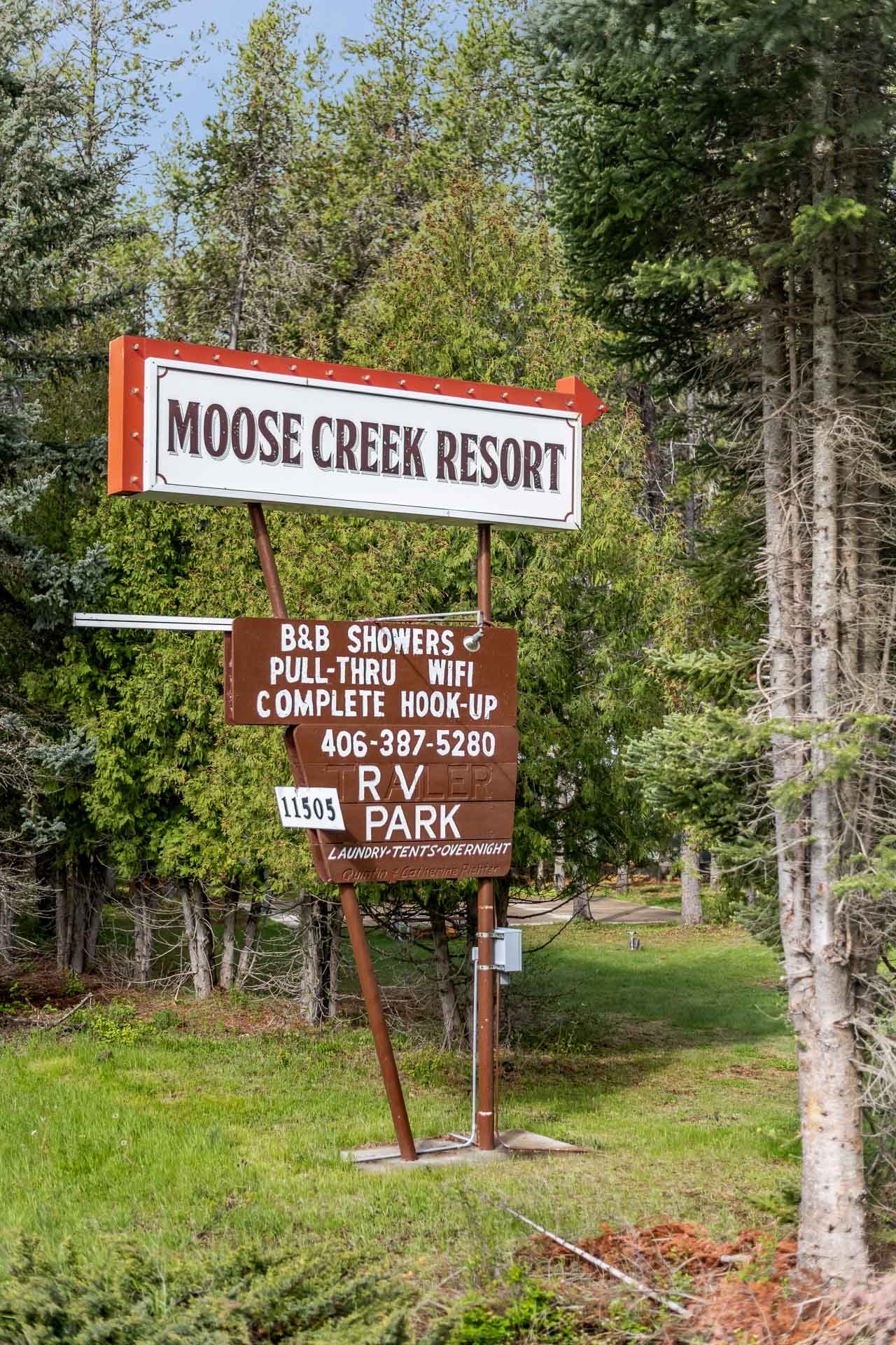 White and orange Moose Creek Resort sign over a brown RV Park panel among evergreen trees near Glacier National Park.