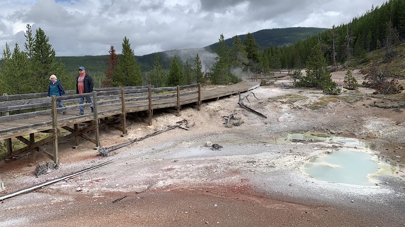 Wooden boardwalk at Artist's Paintpots in Yellowstone National Park, with two hikers, steaming mineral pools.