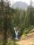 Dense evergreen forest frames a rocky waterfall feeding a pool, with a rugged mountain in Glacier National Park.