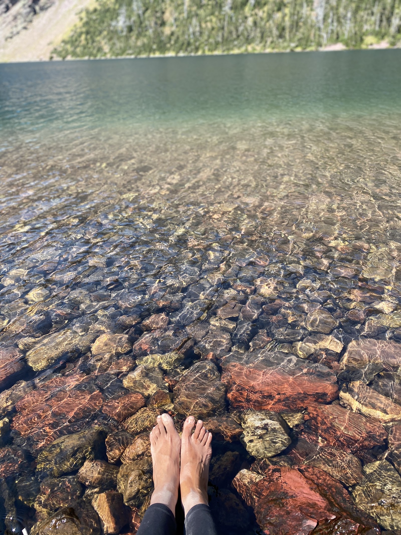 Feet resting on colorful rocks in shallow Otokomi Lake with clear turquoise water and a forested shoreline, Glacier National Park.