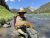 Fisherman along the Yellowstone River in Yellowstone National Park shows off a brown trout with forested hills in the background.