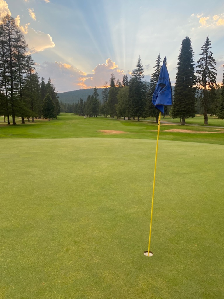 Golf hole with yellow flag on a bright green putting surface near Glacier National Park, framed by tall pines at a lakeside course