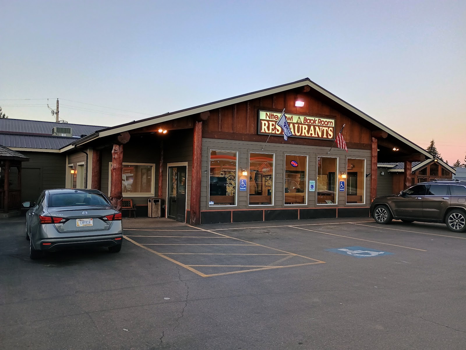 Rustic restaurant building in Glacier National Park at dusk, with wooden beams, large windows, and cars in the parking lot.