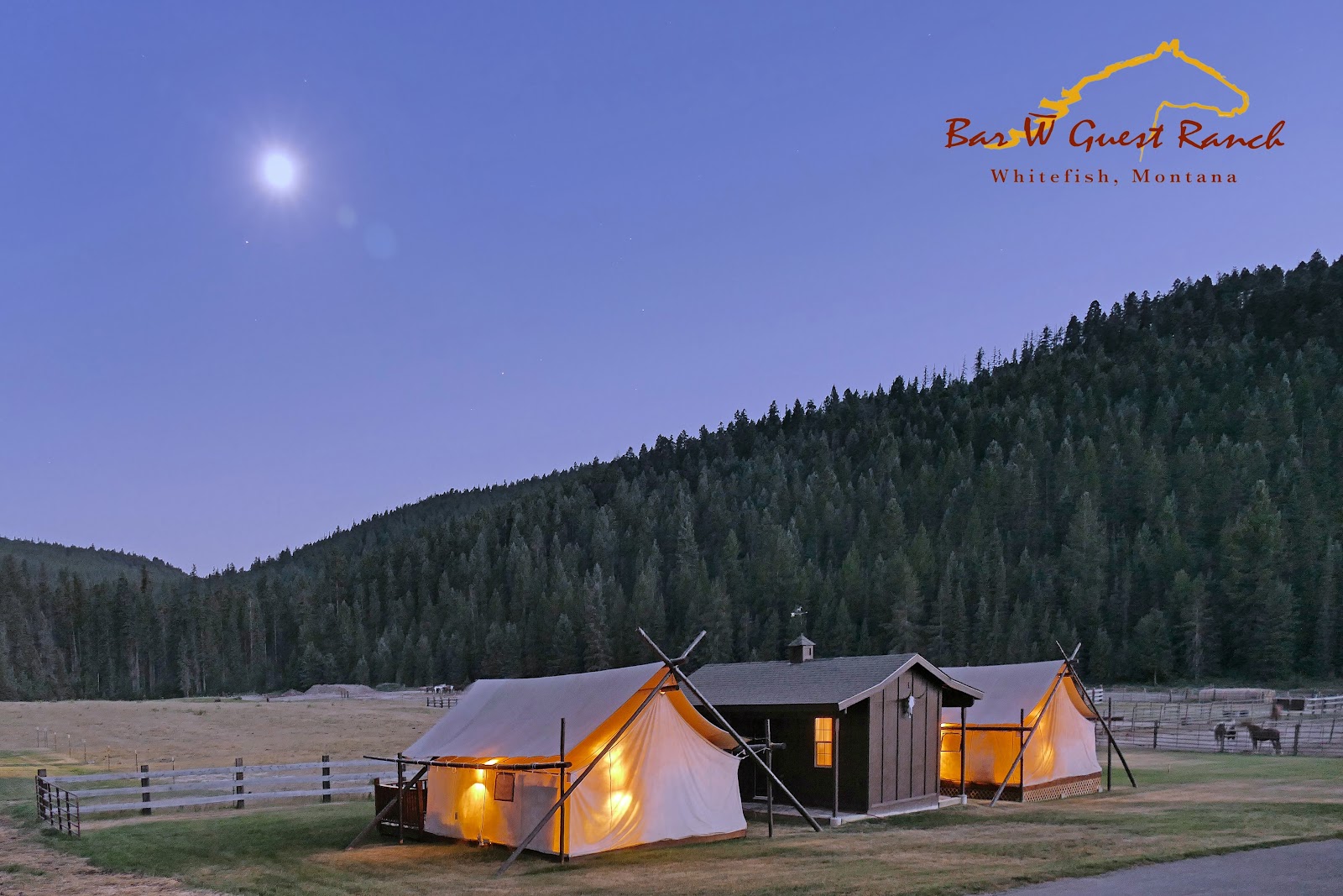 Glacier National Park campsite with lit tents and a central lodge under a moonlit sky at Bar W Guest Ranch campground.