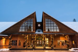 Lodge exterior with twin peaked roofs, warm lights, and snow surrounding Glacier National Park lodgefront at dusk.
