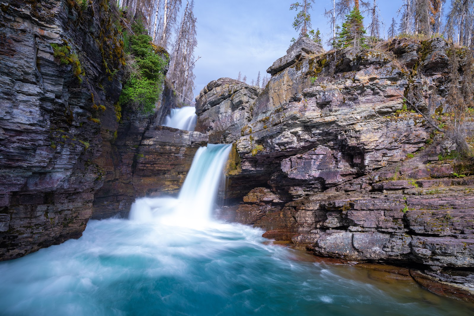 Two-tier waterfall pours over rugged, mossy rock cliffs into a turquoise pool in Glacier National Park.