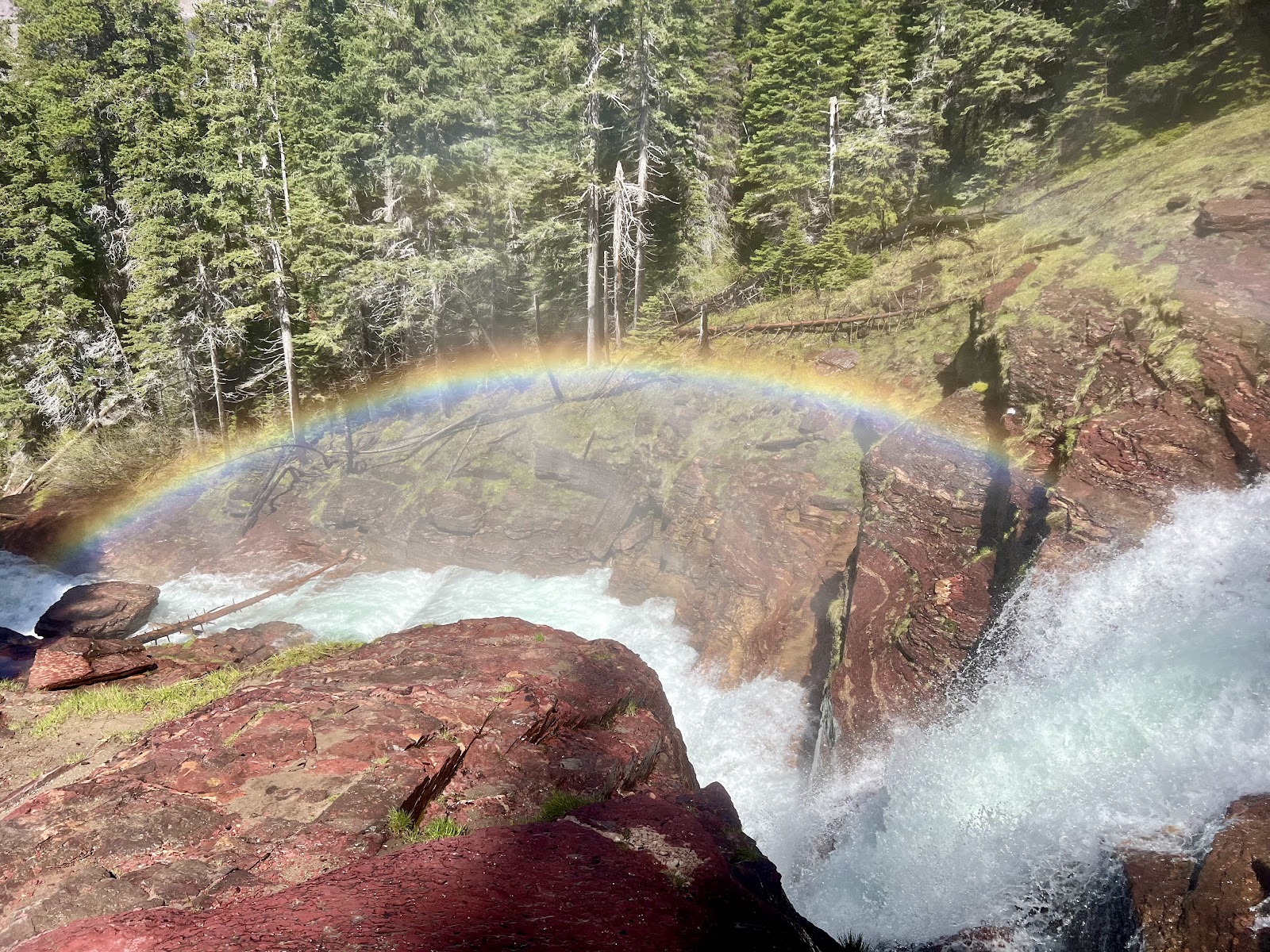 Virginia Falls cascades over red rock cliffs with a rainbow arc over spray in Glacier National Park.