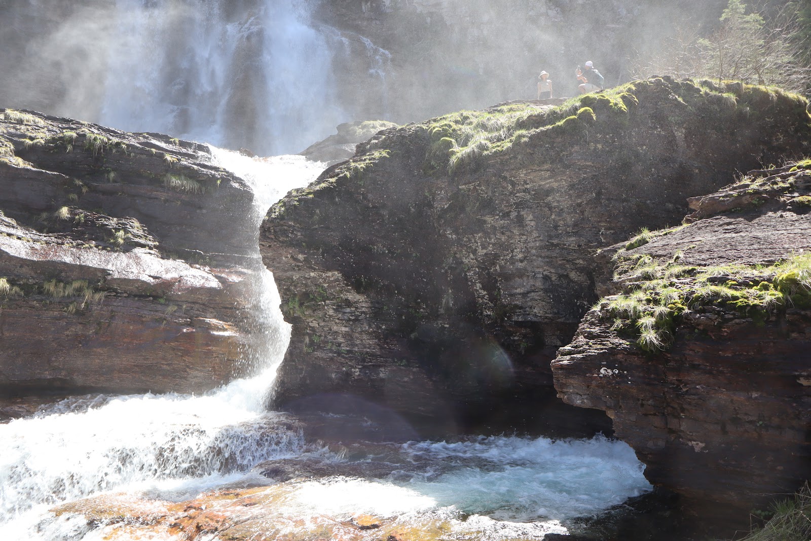 Waterfall spills over layered rocks into a turquoise pool in Glacier National Park, with misty spray and hikers on a grassy ledge above.