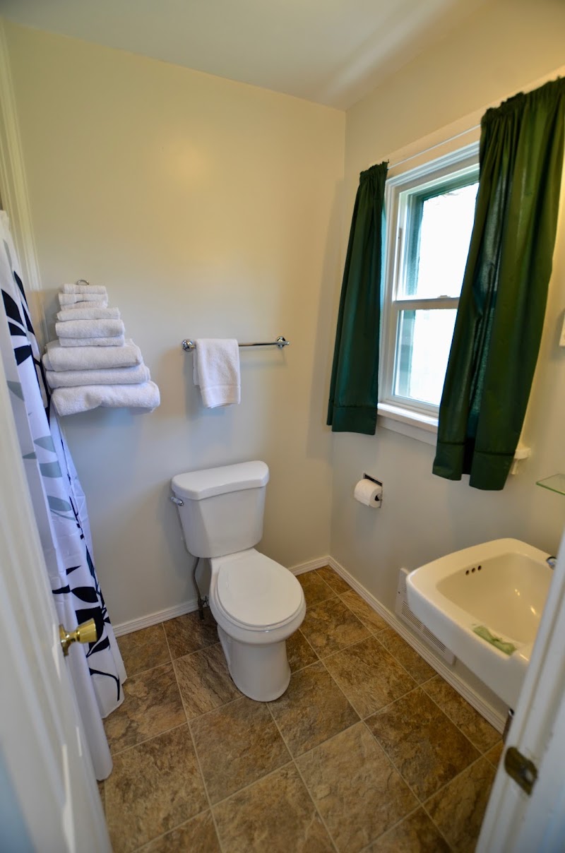 Cozy bathroom in Yellowstone National Park featuring a white toilet, wall towel rack, stacked towels, green-curtained window, and a small sink.