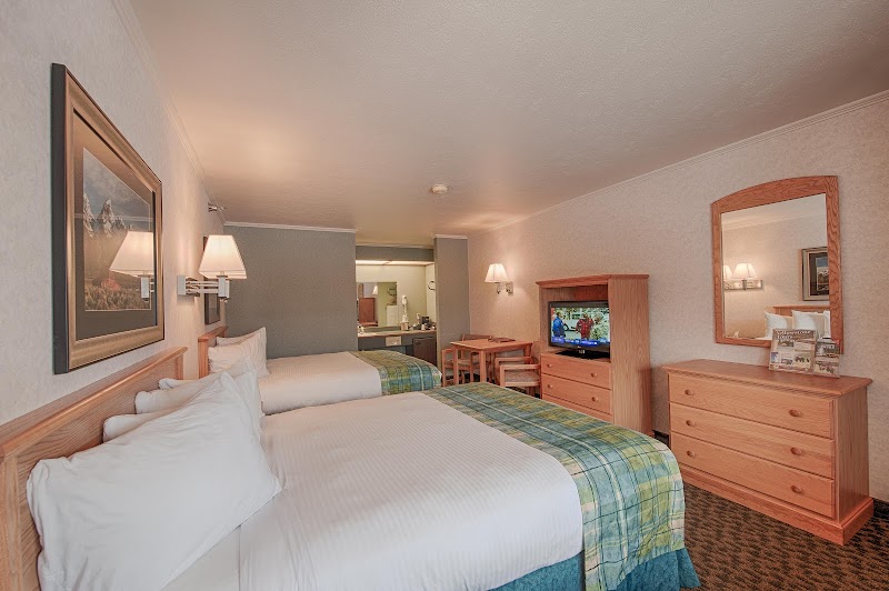 Two queen beds with white linens and green plaid throws, wood dresser, TV, and lamps in a Yellowstone National Park room.