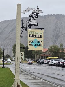 Yellowstone National Park: decorative hanging sign with a cartoon waiter above a wet street by Mammoth Campground, mountains beyond.