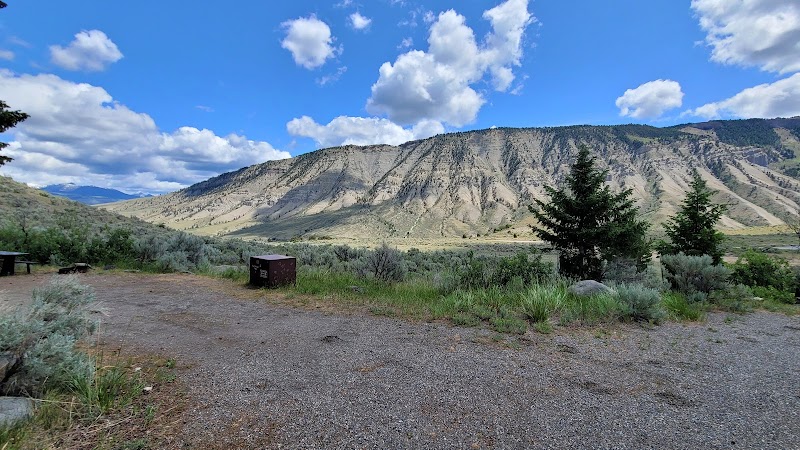 Gravel campground in Yellowstone National Park with a picnic table, bear box, sagebrush, pines, and rugged ridged mountains in the distance.