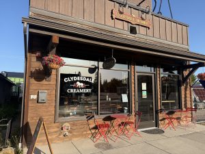 Rustic wooden storefront with hanging flower baskets and red outdoor tables outside Glacier National Park’s creamery storefront.