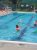 Swimmers in a bright outdoor pool at a Glacier National Park lodging area, with a metal ladder, green shade awning, and fenced surroundings.