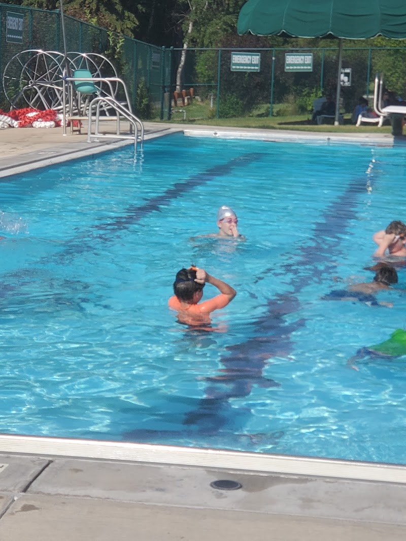 Swimmers in a bright outdoor pool at a Glacier National Park lodging area, with a metal ladder, green shade awning, and fenced surroundings.