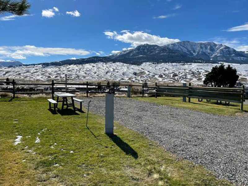 Grassy overlook with a wooden picnic table, benches, and a gravel path, facing snow-covered Yellowstone National Park mountains.