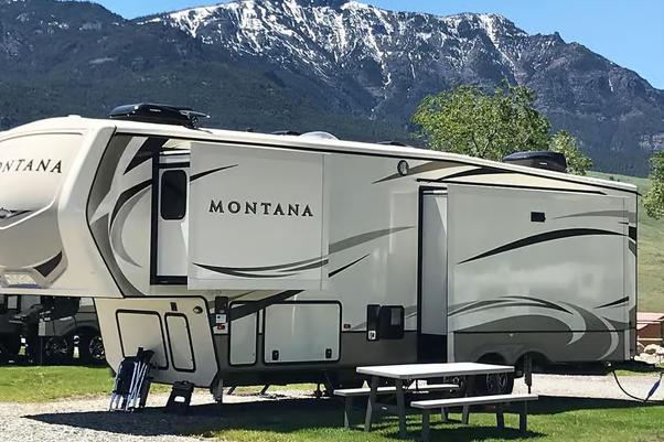 Large beige fifth-wheel trailer in a Yellowstone National Park campground with a picnic table and snow-capped mountains.