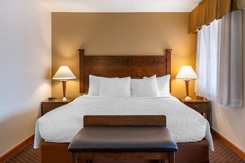 A warmly lit hotel room with a large wooden headboard bed, white bedding, twin nightstands with lamps, a bench at the foot, and a window with beige curtains in Yellowstone National Park.